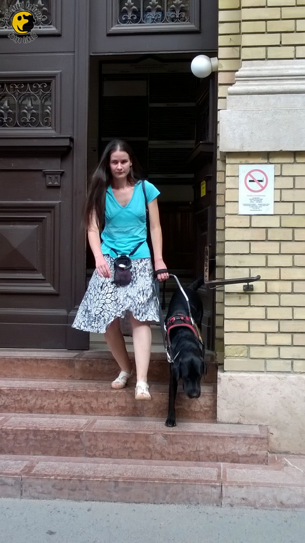 Éppen egy épületből lépnek ki, a lépcsőkön haladnak. Anikón elegáns kék felső és szoknya van